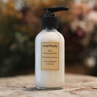 Front view of Rose Cleansing Milk bottle with pump, showing evanhealy label and creamy white cleanser on a blurred floral background