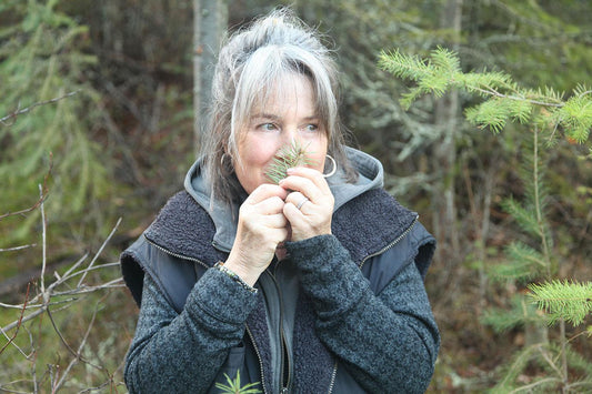 Woman smelling a Douglas fir sprig in a forest, wearing a gray hooded jacket