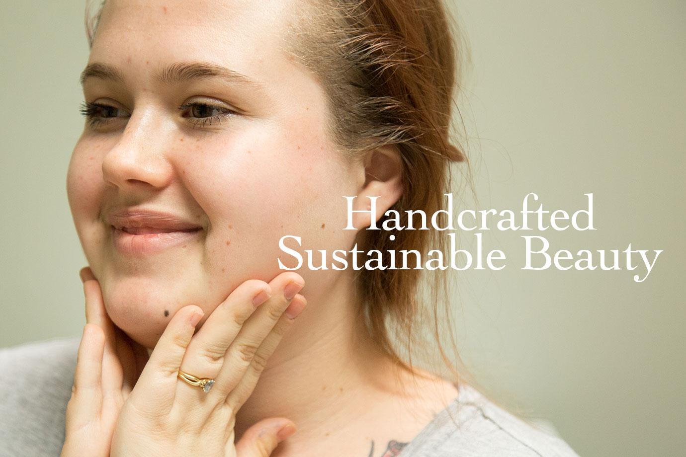 Woman touching her cheeks and smiling in a softly lit setting with overlaid text ‘Handcrafted Sustainable Beauty’