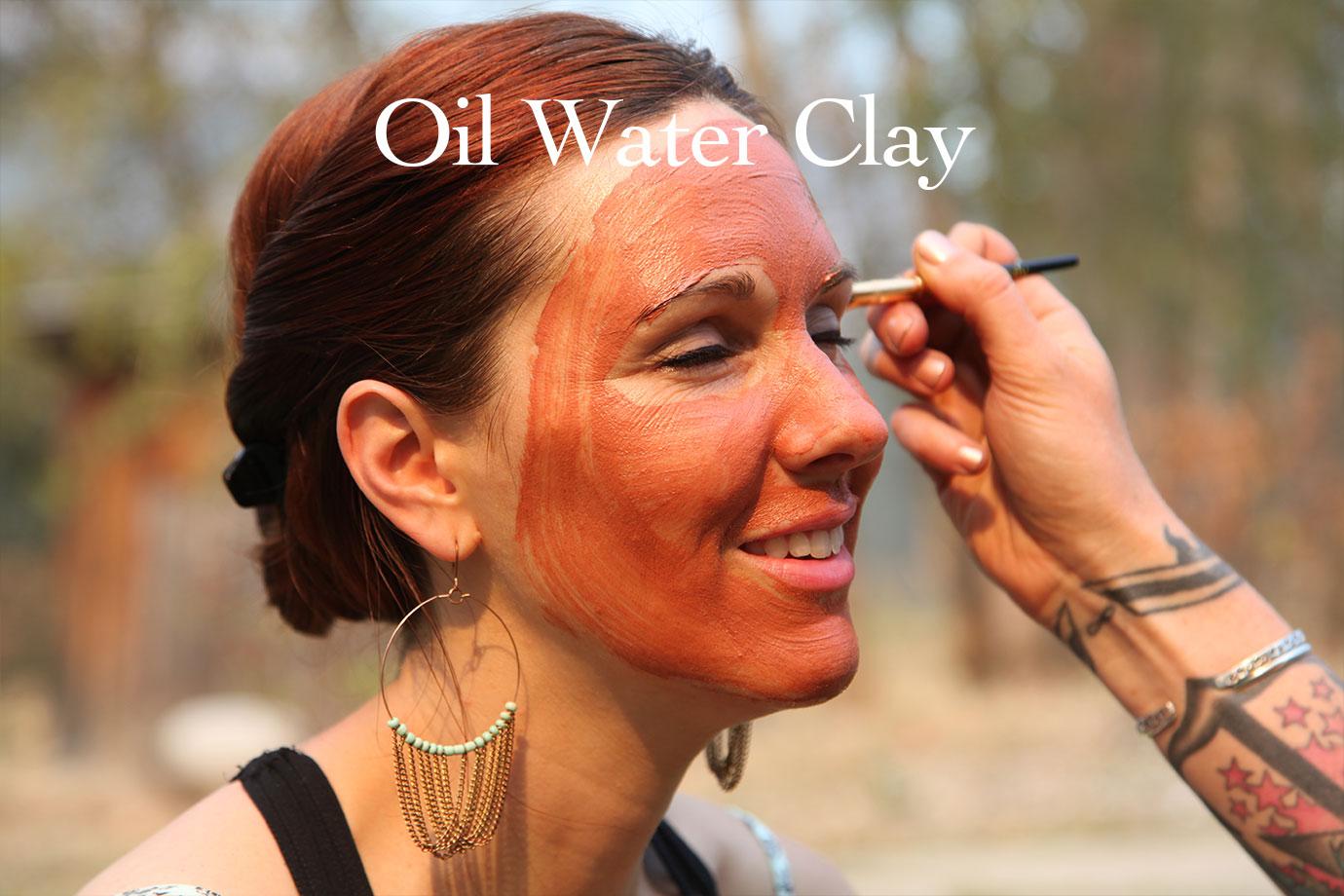 Woman smiling as a cosmetician applies a red clay face mask outdoors, overlay text Oil Water Clay