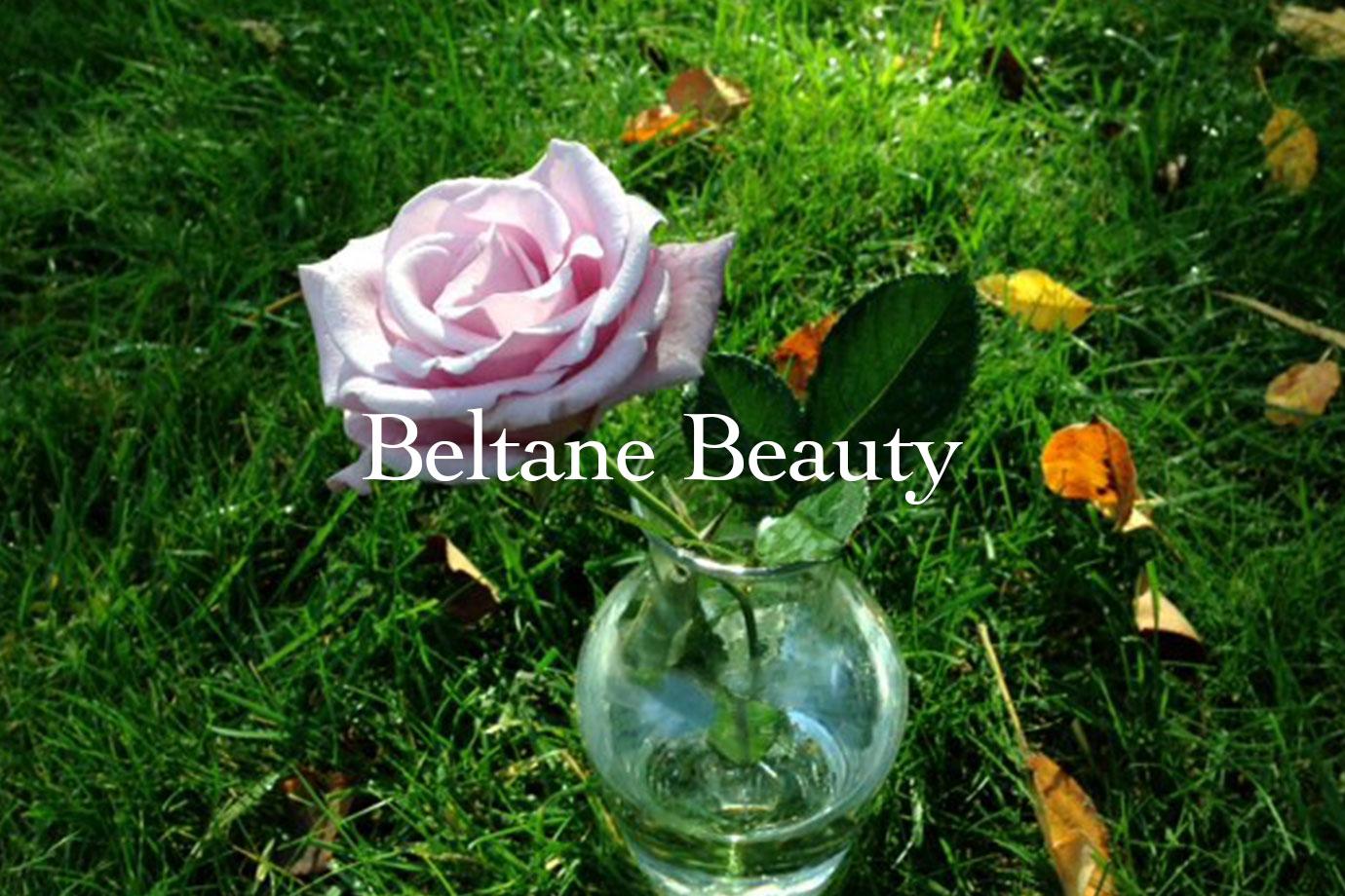 Pink rose in a small glass vase on green grass, overlaid with the headline Beltane Beauty