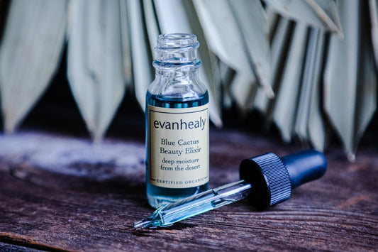 Front view of Blue Cactus Beauty Elixir bottle showcasing blue serum with glass dropper on wooden surface and blurred palm-leaf background