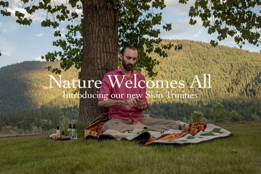 Man sitting cross-legged on a blanket under a tree applying skincare, bottles beside him and forested hills behind