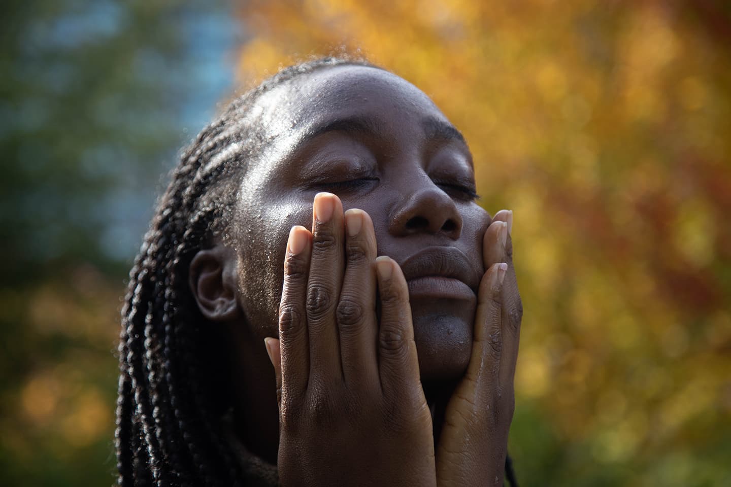 Close-up of a person with braided hair gently pressing moisturized cheeks with eyes closed against a blurred autumn foliage background