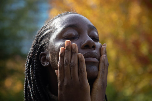 Close-up of a person with braided hair gently pressing moisturized cheeks with eyes closed against a blurred autumn foliage background