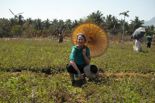 Woman crouching in a field wearing a teal top, holding a yellow paper parasol and a hat, with palm trees and people in the background