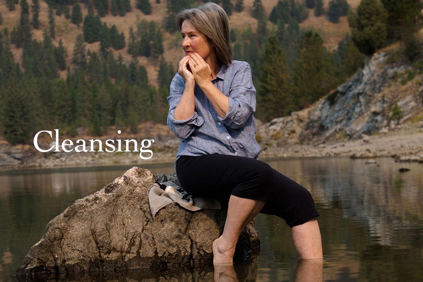 Woman sitting on a rock at a mountain lake, dipping her feet in the water with overlaid headline “Cleansing”