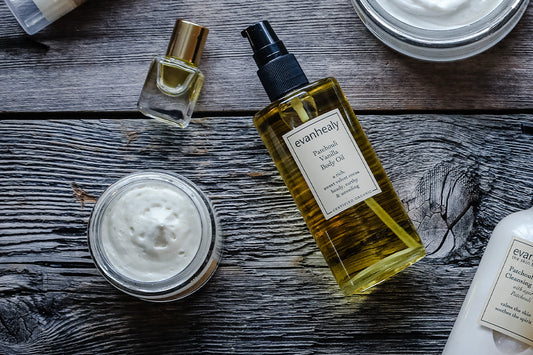 Evanhealy patchouli products on weathered wood: Patchouli Vanilla Body Oil, open cream jar, and a small perfume bottle