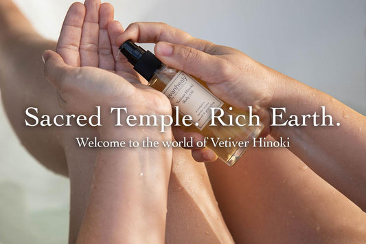 Hands dispensing Vetiver Hinoki body oil into a cupped palm over bare legs in soft natural light