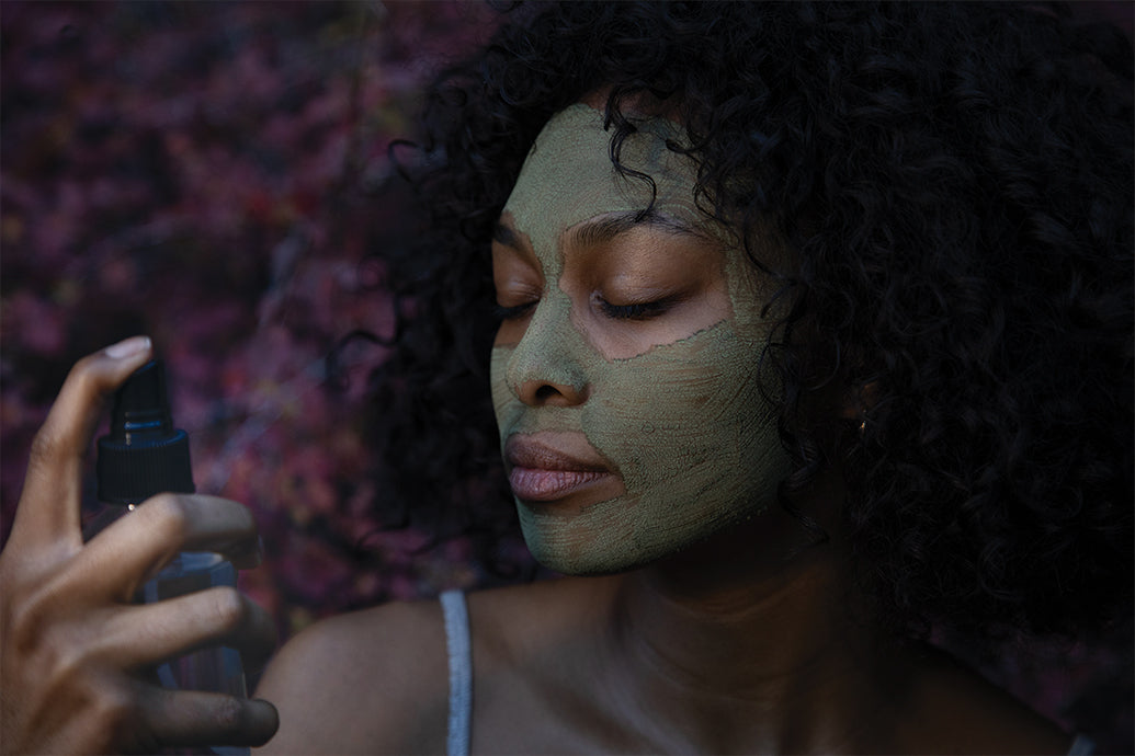 Woman with green facial mask spraying facial mist with eyes closed against blurred purple foliage background