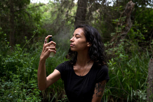 Woman spraying facial mist toward her face while seated in a shaded forest