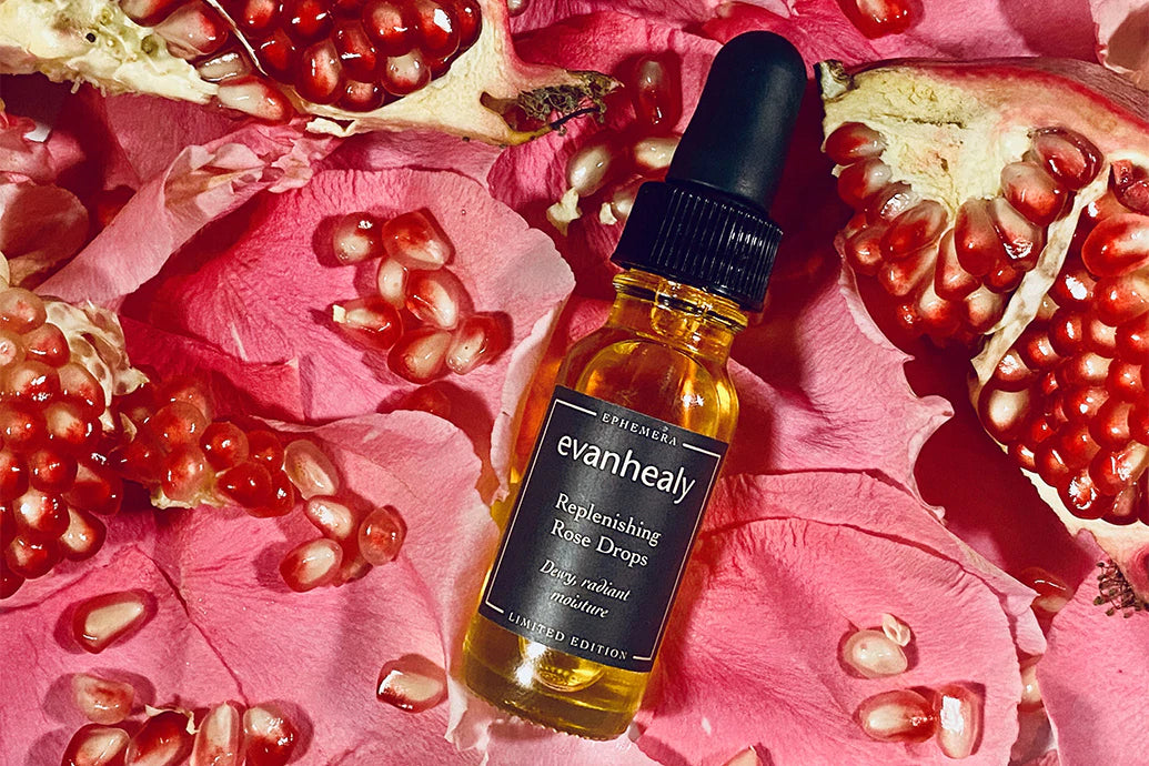 Top down view of evanhealy Replenishing Rose Drops bottle with amber oil on pink rose petals and pomegranate seeds
