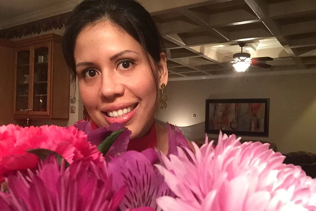 Woman smiling at camera behind a bouquet of pink and purple flowers in a warmly lit living room