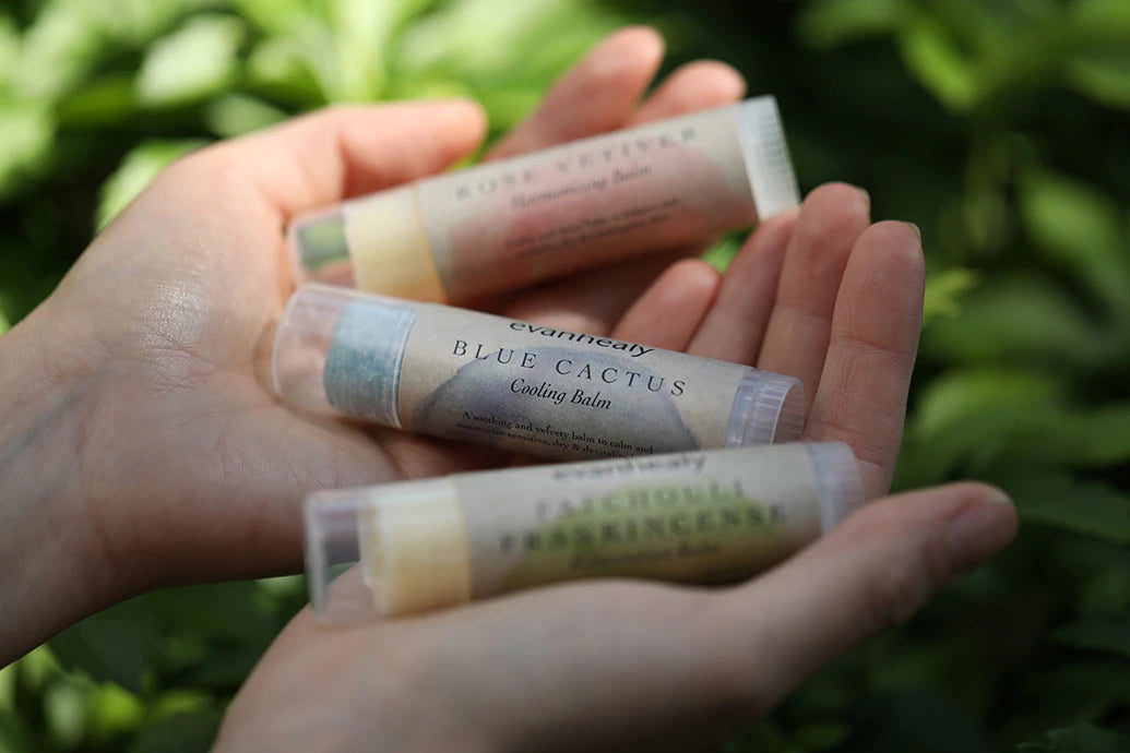 Close-up of three face balm tubes in cupped hands showing twist-up tubes and paper labels against blurred green foliage