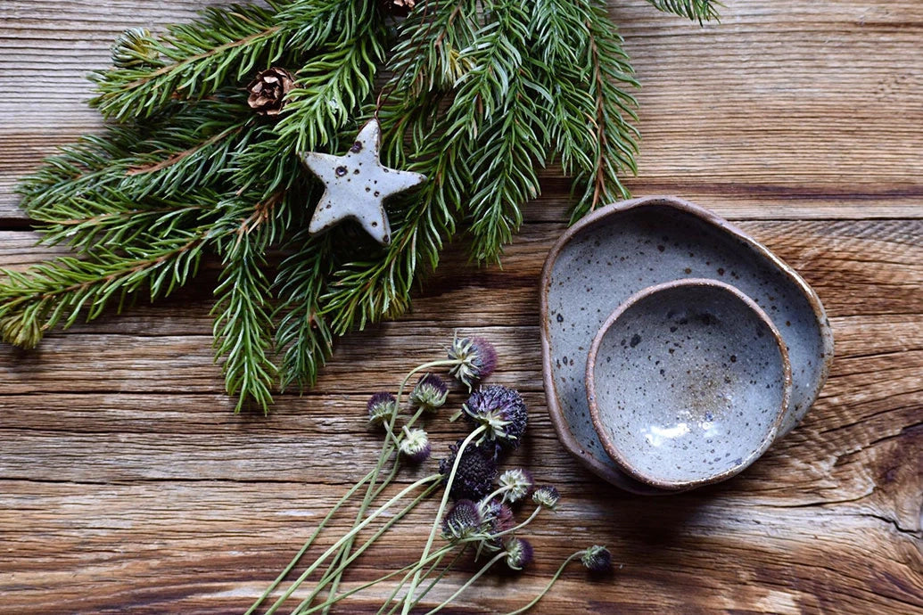 Evergreen sprig and star ornament beside stacked handmade ceramic bowls and dried thistle flowers on a rustic wooden table