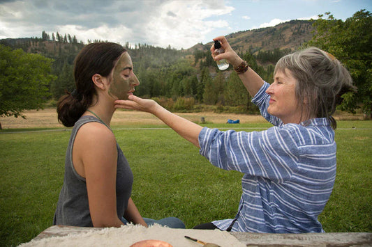 Older woman spraying facial mist on younger woman wearing a green clay face mask while they sit on grass with tree-covered hills behind them