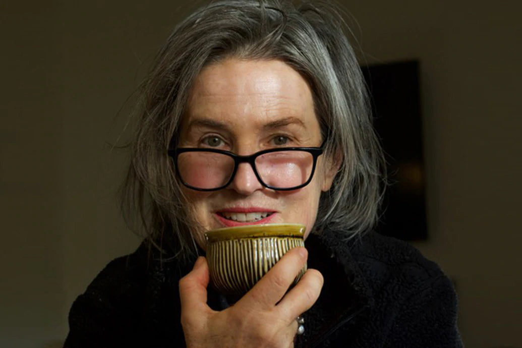 Person holding a striped ceramic teacup to their mouth, wearing glasses and a dark jacket against a dim background