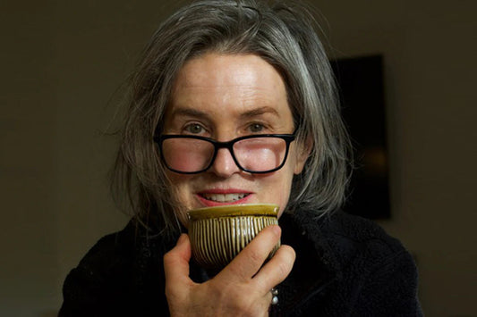 Person holding a striped ceramic teacup to their mouth, wearing glasses and a dark jacket against a dim background