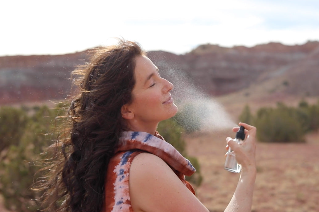 Woman with long dark hair spraying facial mist onto her face with eyes closed in a sunlit desert landscape