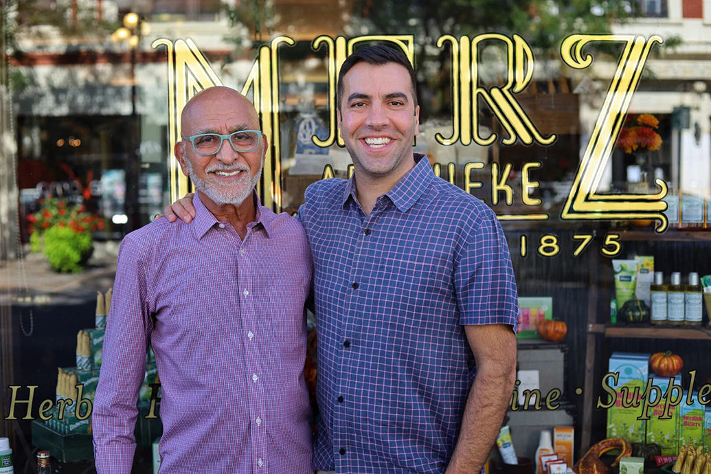 Two men smiling and posing in front of a Merz Apothecary storefront window with gold lettering