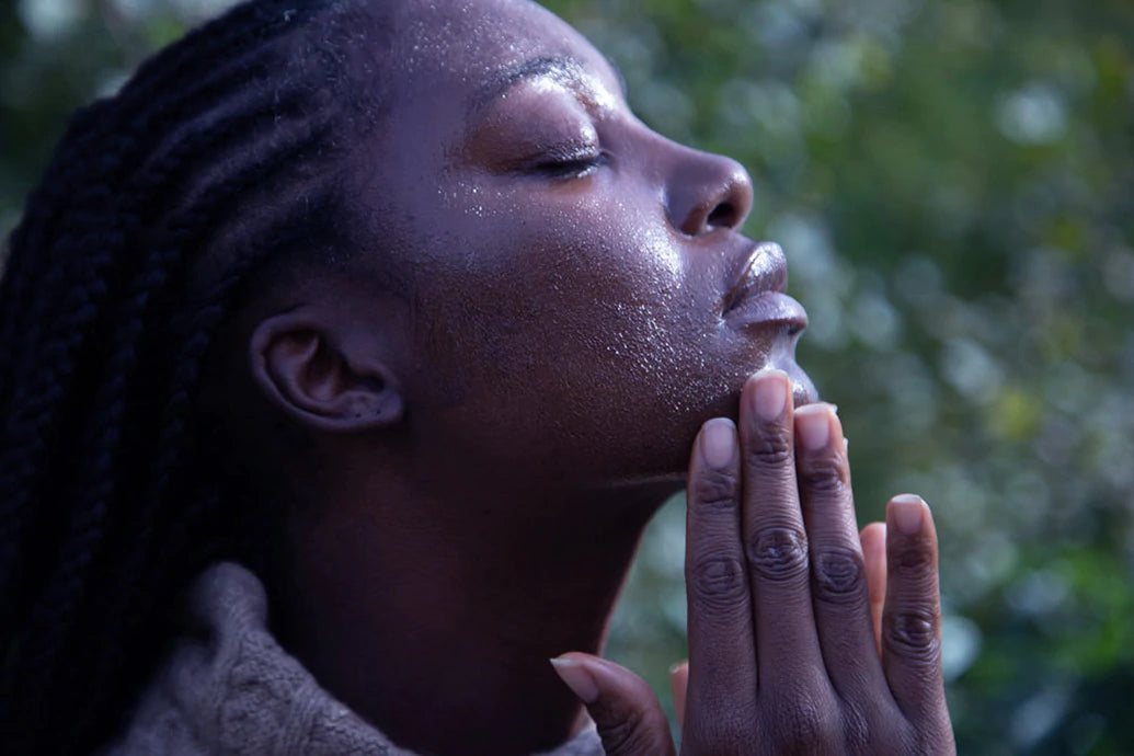 Woman with shea butter on her face, eyes closed with hands at her chin against a blurred green background