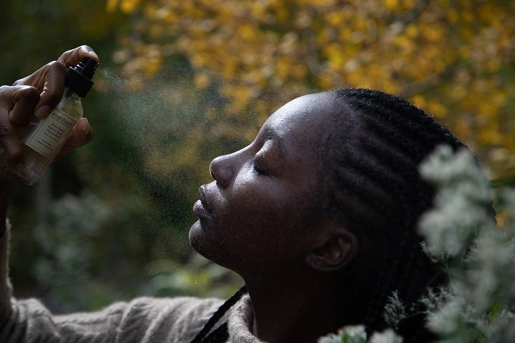 Woman with braided hair spraying hydrosol mist onto her face in a leafy outdoor setting