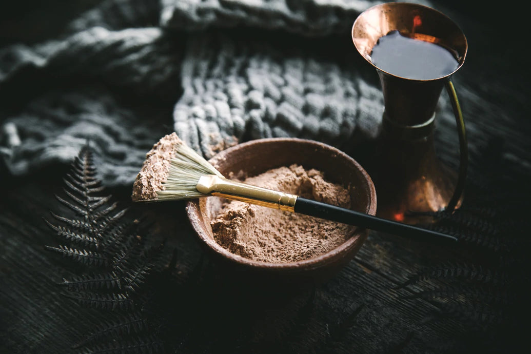 Powdered clay in a small bowl with a brush coated in clay, a copper water pitcher and a knitted cloth on a dark surface