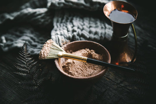 Powdered clay in a small bowl with a brush coated in clay, a copper water pitcher and a knitted cloth on a dark surface