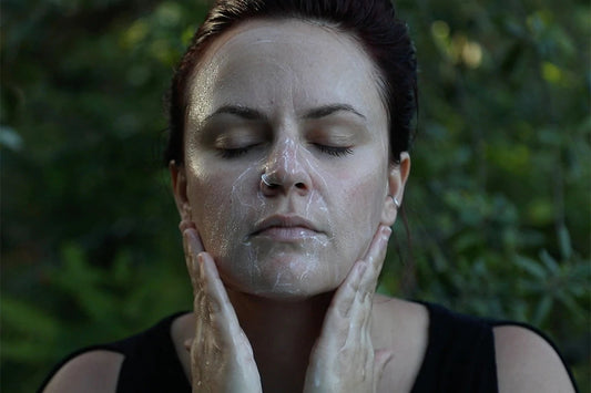 Woman cleansing her face with foamy cleanser, eyes closed and hands on her jaw, blurred green foliage background