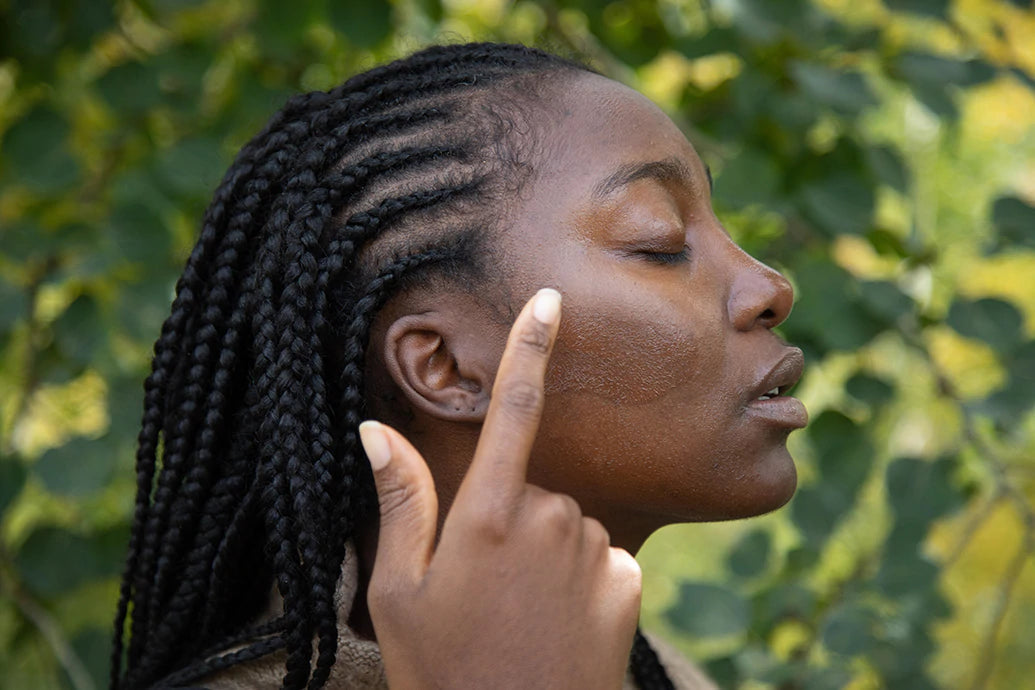 Close-up of a woman with braided hair applying moisturizer to her cheek outdoors against a leafy background