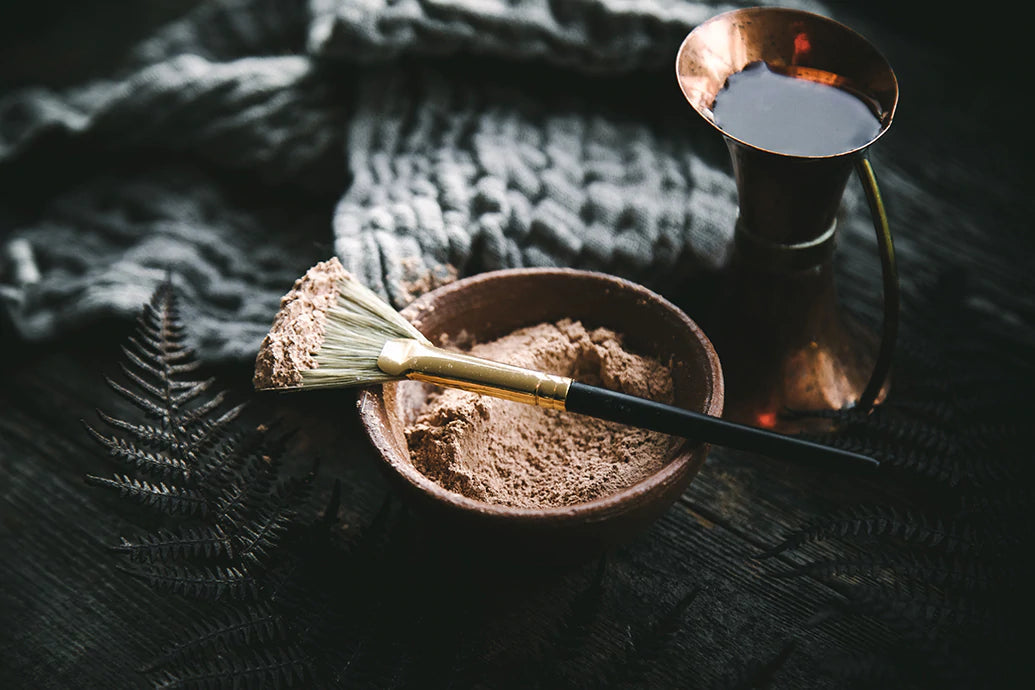 Bowl of powdered clay with a flat mask brush and copper pitcher on a dark wooden table with a knit cloth