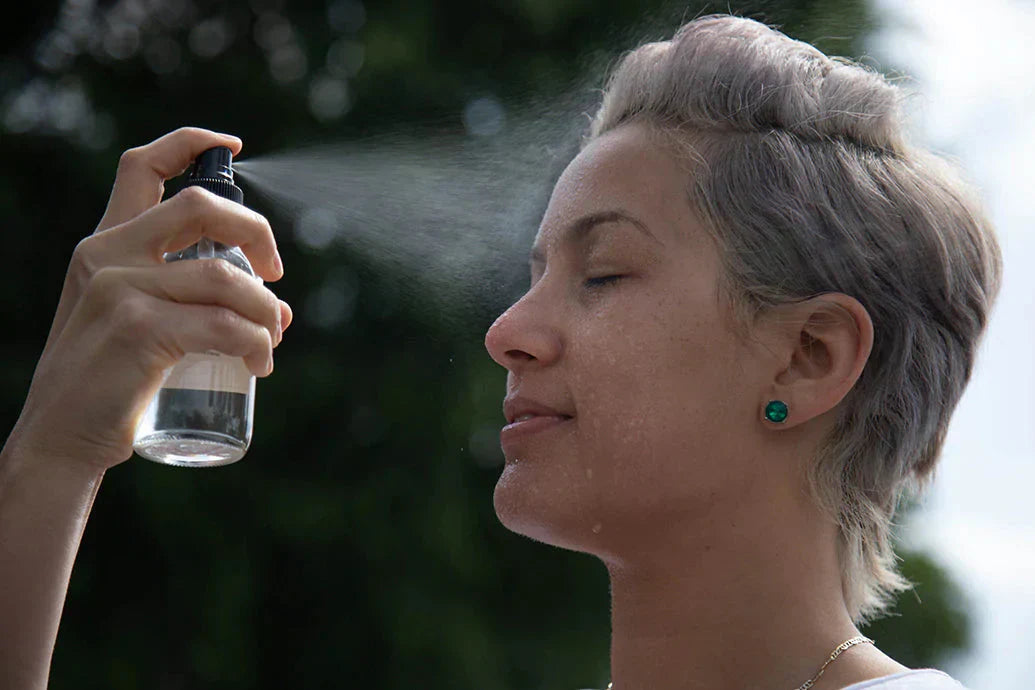 Person spraying facial mist onto their face with eyes closed outdoors against a blurred green background