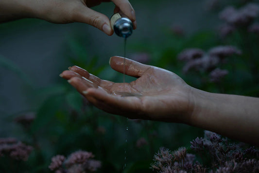 Hands pouring facial oil from a small bottle into an open palm over pink garden flowers