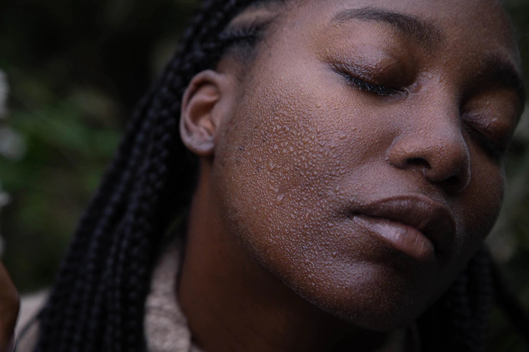 Woman with braided hair closing her eyes while water beads on her cheek against a blurred outdoor background