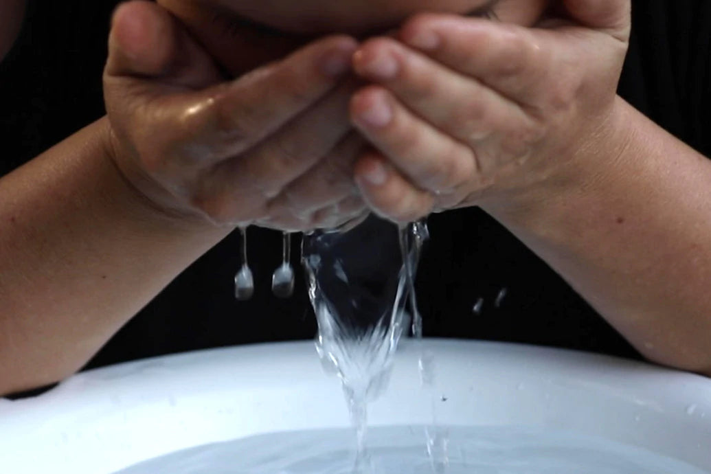 Person splashing water onto their face while cupping water over a white basin with dark background