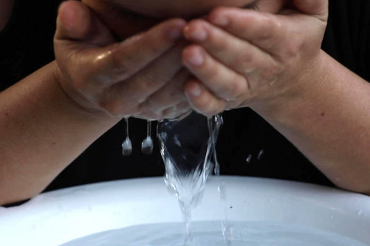 Person splashing water onto their face while cupping water over a white basin with dark background