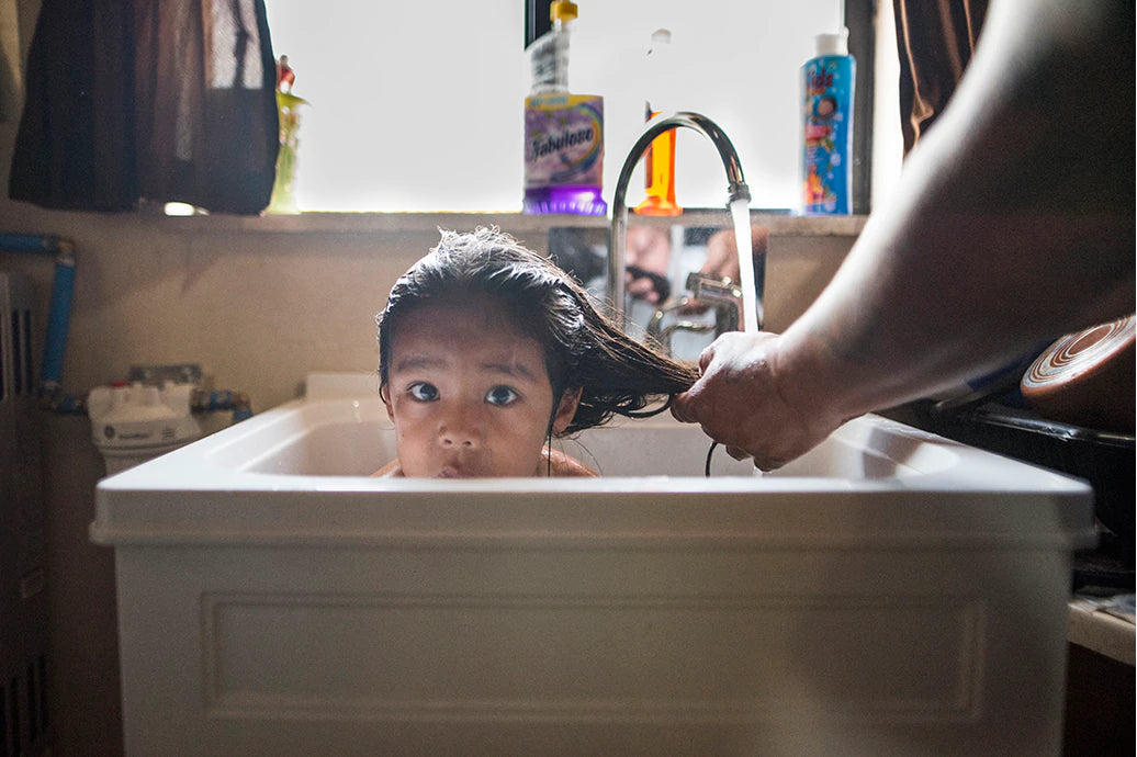 Child having hair washed in a white utility sink as an adult rinses hair under a faucet by a window