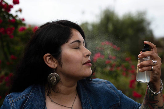Woman spraying facial hydrosol toward her face outdoors in a garden with blurred red flowers