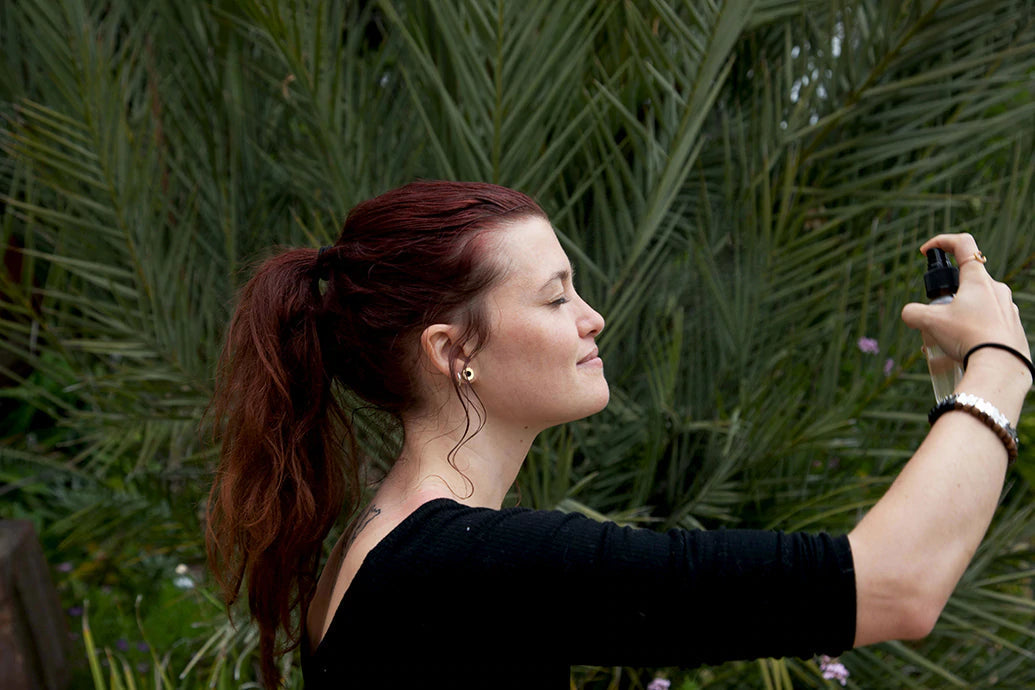 Woman with a ponytail spraying a mist from a spray bottle outdoors in a garden with palm leaves in the background