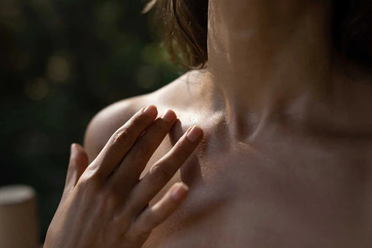 Close-up of a woman’s hand touching her sunlit shoulder and collarbone with a blurred green background