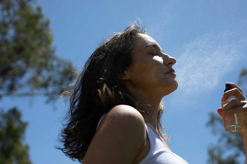 Woman spraying mist onto her face from a small bottle outdoors against a blue sky