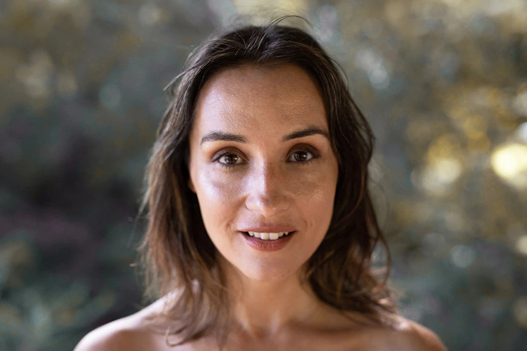 Woman smiling at camera outdoors with dewy skin and soft autumn-toned bokeh background