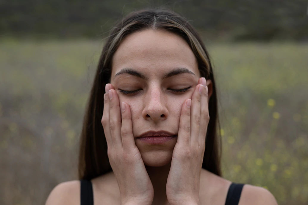 Young woman gently pressing her palms to her cheeks with eyes closed in a blurred meadow background