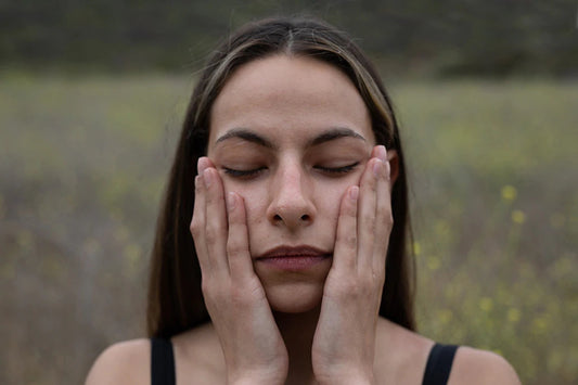 Young woman gently pressing her palms to her cheeks with eyes closed in a blurred meadow background