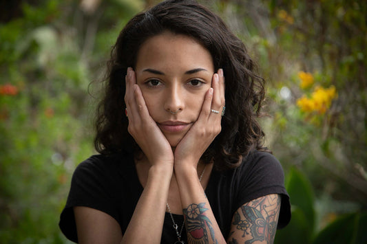 Young woman resting her face in her hands, showing floral forearm tattoos and a septum ring against a blurred garden background