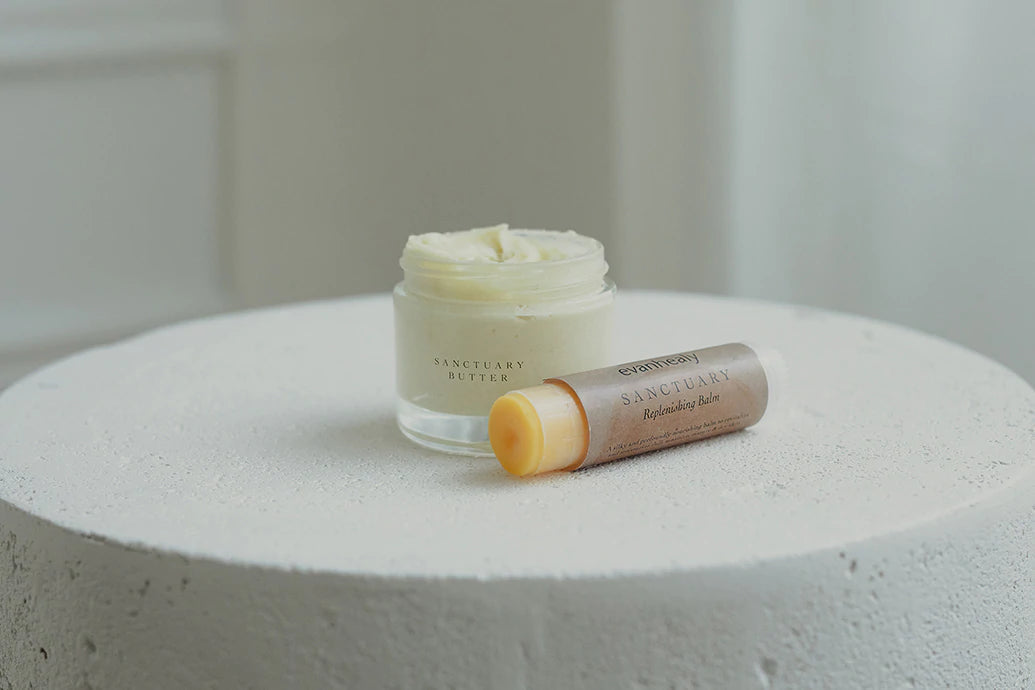 Front view of Sanctuary Butter jar and Sanctuary Replenishing Balm stick showing creamy butter texture and balm tip on a round white textured pedestal