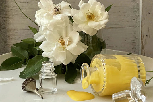 Glass jar of yellow liquid balm spilled on a white tabletop beside a small glass vial and white gardenia flowers in a vase