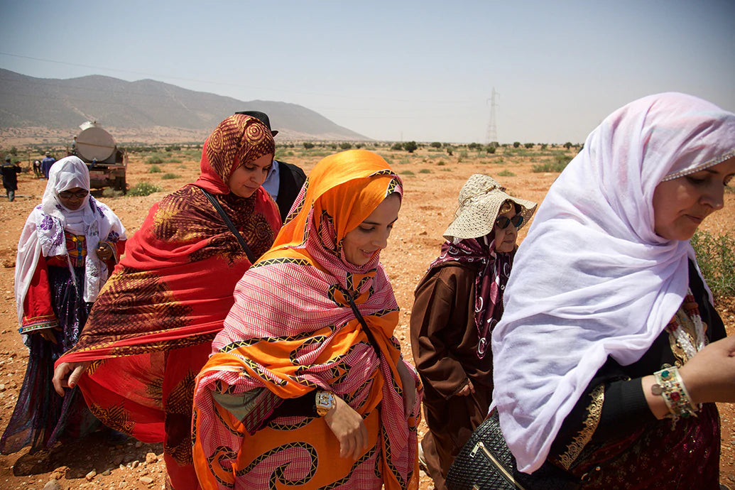 Group of women in colorful headscarves walking across a dry field with mountains and sparse trees in the distance