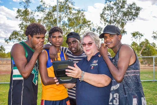 Woman holding a tablet and showing it to four young men standing together in a grassy park with trees and a chain-link fence