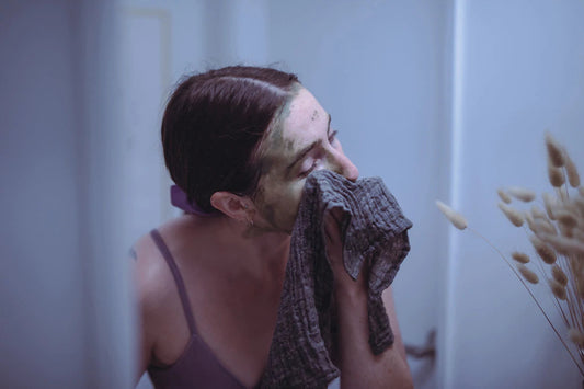 Woman removing a green facial mask with a cloth while looking into a softly lit bathroom mirror